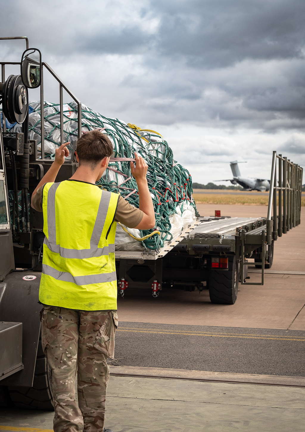 A military forked vehicle loads cargo on a netted pallet onto a Condec loader on an airfield. A member of 1 Air Mobility Wing, in camouflage trousers and a high-visibility vest, guides The driver of the vehicle, enabling a safe movement of the pallet onto the Condec. there is an Atlas (A400M) aircraft visible in the background under a cloudy sky.
